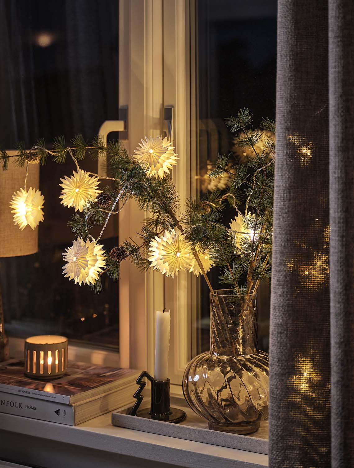 Bir vazoda duran çam dallarını saran yıldız şeklindeki ışık zincirleri, gece vakti pencere kenarına ışıltı katıyor.