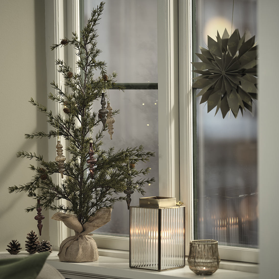 FENRIR yılbaşı ağacı, LUCIA süslemeleri, UVIT fener, SAFRAN tealight mumluk ve HRYM yılbaşı yıldızı Tealight mumluk, fener ve süslemelerle donatılmış yapay yılbaşı ağacı pencere kenarında.